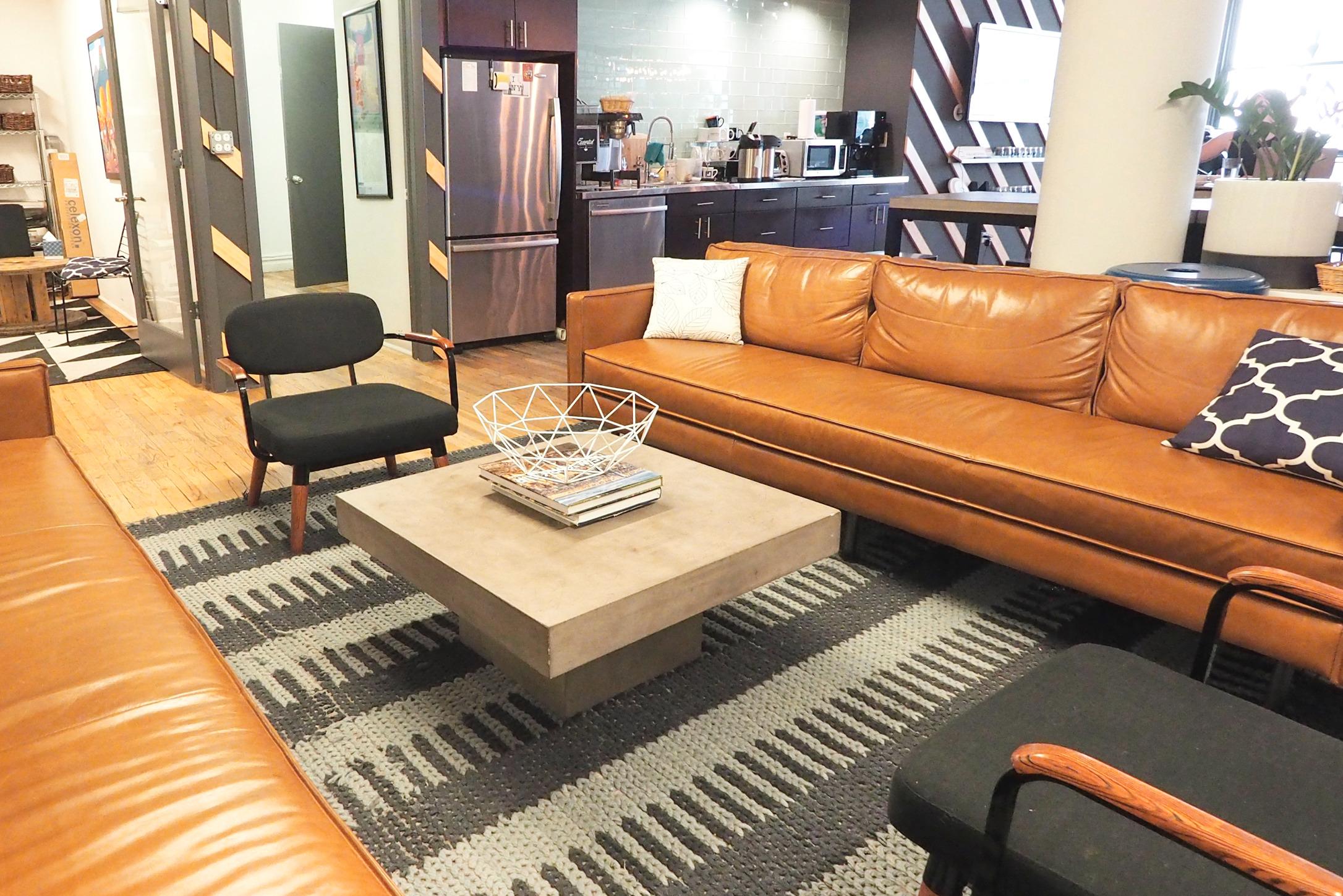 An office lounge area with long leather couch, coffee table, and kitchen in the background